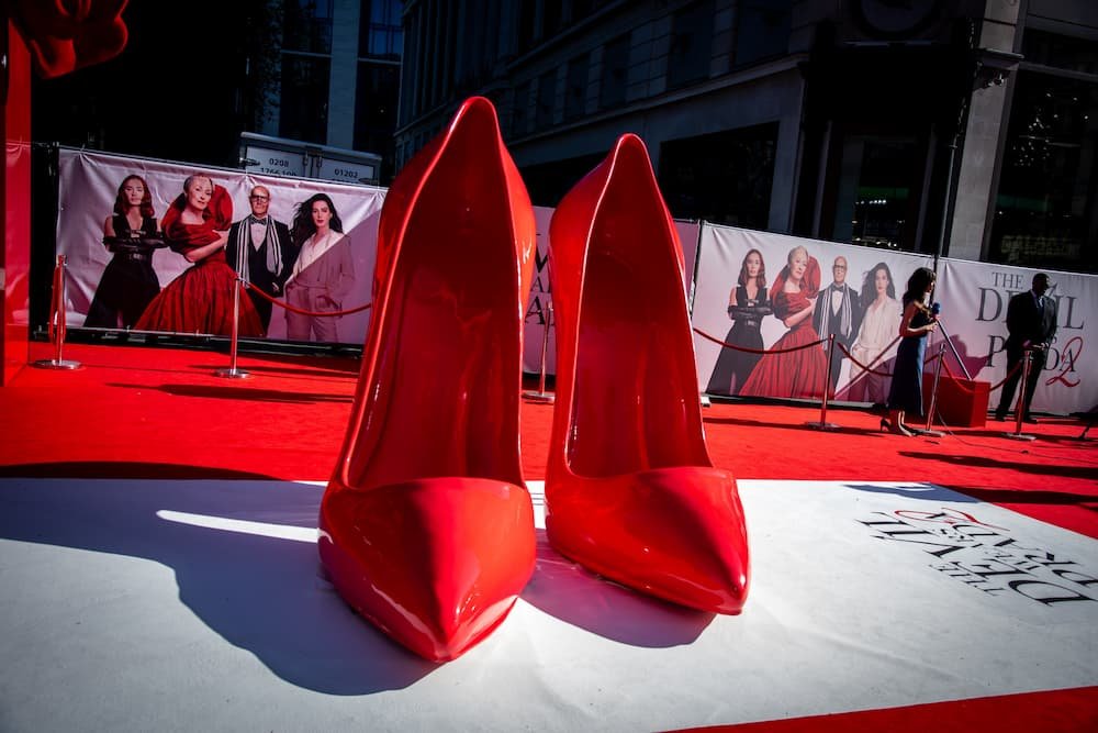 Red stiletto heels on a red carpet with a fashion-themed backdrop inspired by The Devil Wears Prada